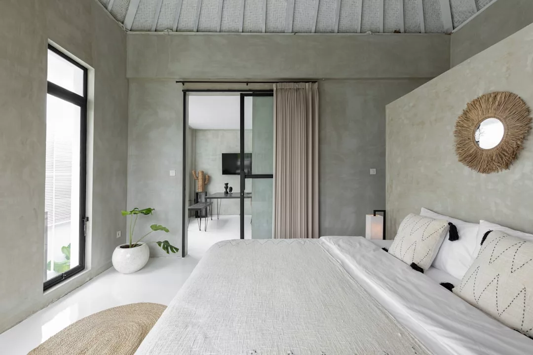 Minimalistisches Schlafzimmer im Mallorca-Stil mit großem Bett, neutralen Farbtönen, dekorativen Kissen, rundem Wandspiegel, Topfpflanze und Glasschiebetüren, die in ein anderes Zimmer führen.