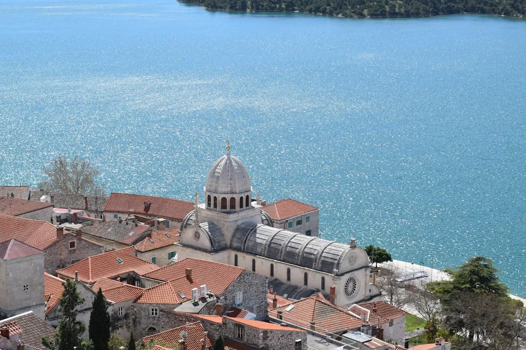Eine steinerne Kathedrale mit einer großen Kuppel überblickt Mallorcas Gebäude mit ihren roten Ziegeldächern entlang der Uferpromenade, mit blauem Wasser und einer grünen Insel im Hintergrund.