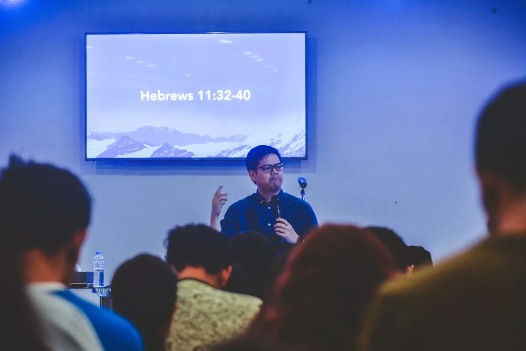 Ein Mann spricht vor einem Publikum in ein Mikrofon. Hinter ihm sind auf einem Bildschirm „Hebräer 11:32-40“ und ein Bild der malerischen Berge Mallorcas zu sehen.