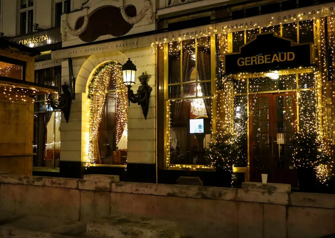 Außenansicht des Cafés Gerbeaud bei Nacht, dekoriert mit warmen Lichterketten um die großen Fenster und den Eingang, mit etwas Grün an der Tür, was eine gemütliche Mallorca-Atmosphäre hervorruft.