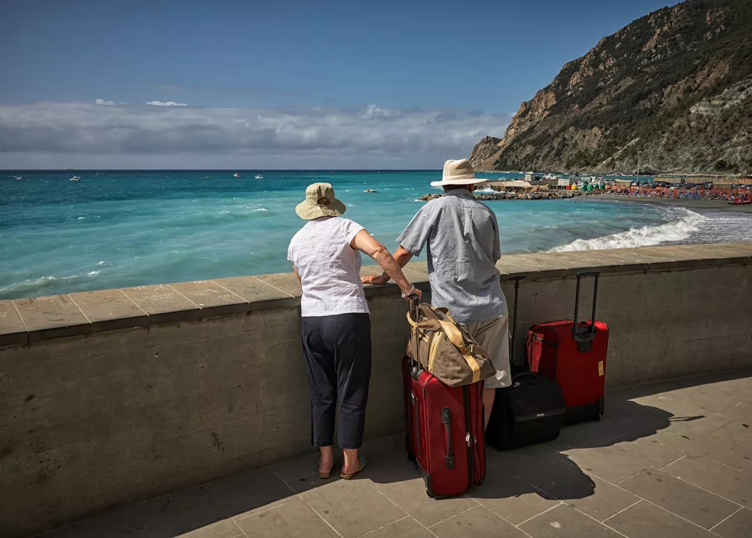 Zwei Menschen mit Hüten und Gepäck stehen an einer Strandpromenade auf Mallorca und blicken auf das türkisfarbene Meer, die felsige Küste und einen überfüllten Strand in der Ferne.