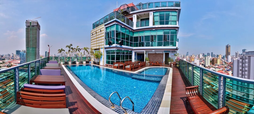 Der Swimmingpool auf dem Dach mit hölzernen Liegestühlen, umgeben von Glasgeländern, bietet einen atemberaubenden Blick auf die Skyline einer modernen Stadt unter klarem Himmel und bringt einen Hauch mallorquinischer Entspannung in die urbane Umgebung.