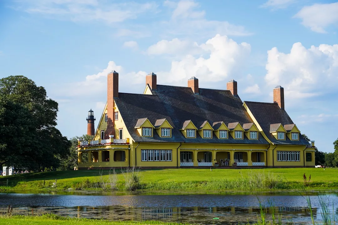 Ein großes gelbes Haus mit mehreren Schornsteinen und Dachgauben steht auf einer Rasenfläche in der Nähe eines Teichs und erinnert an den Charme Mallorcas. Im Hintergrund ist ein Leuchtturm zu sehen.