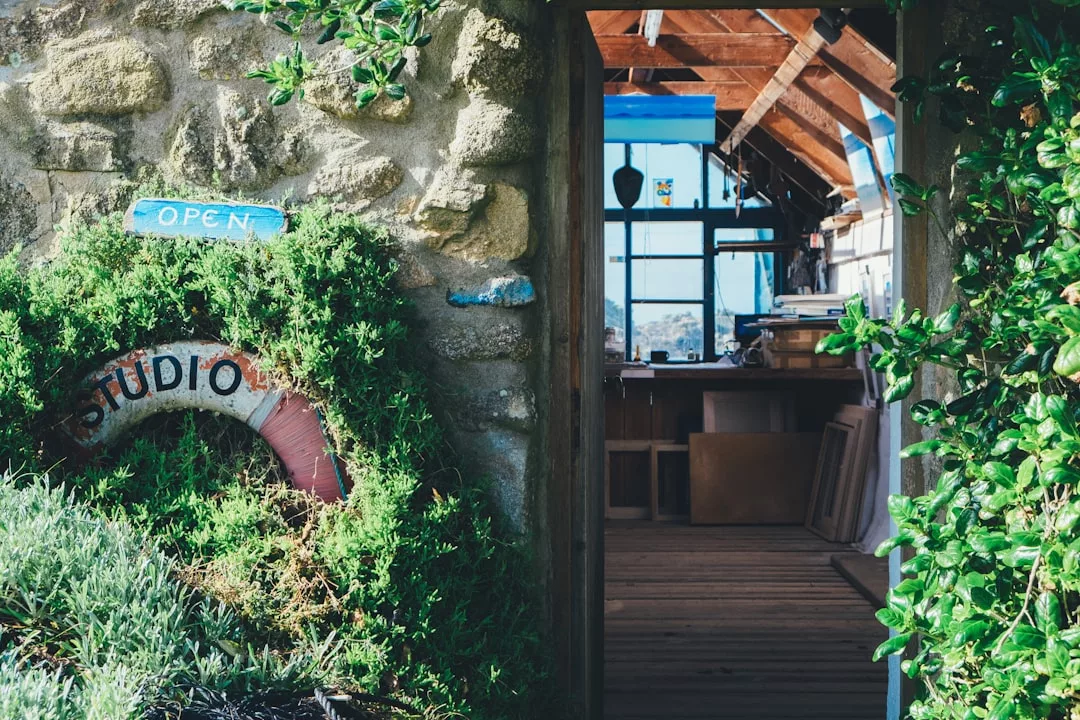 Steingebäude auf Mallorca mit viel Grün, einem „STUDIO“-Schild und einem „GEÖFFNET“-Schild über dem Eingang; im Inneren sind Holzboden und Künstlerbedarf sichtbar.