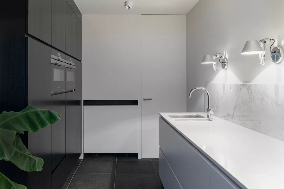 Minimalistische Küche im Mallorca-Stil mit weißen Arbeitsplatten, schwarzen Schränken, eingebautem Ofen, modernem Spülbecken, Marmorrückwand, Wandleuchten und einer großen grünen Pflanze in der Ecke.