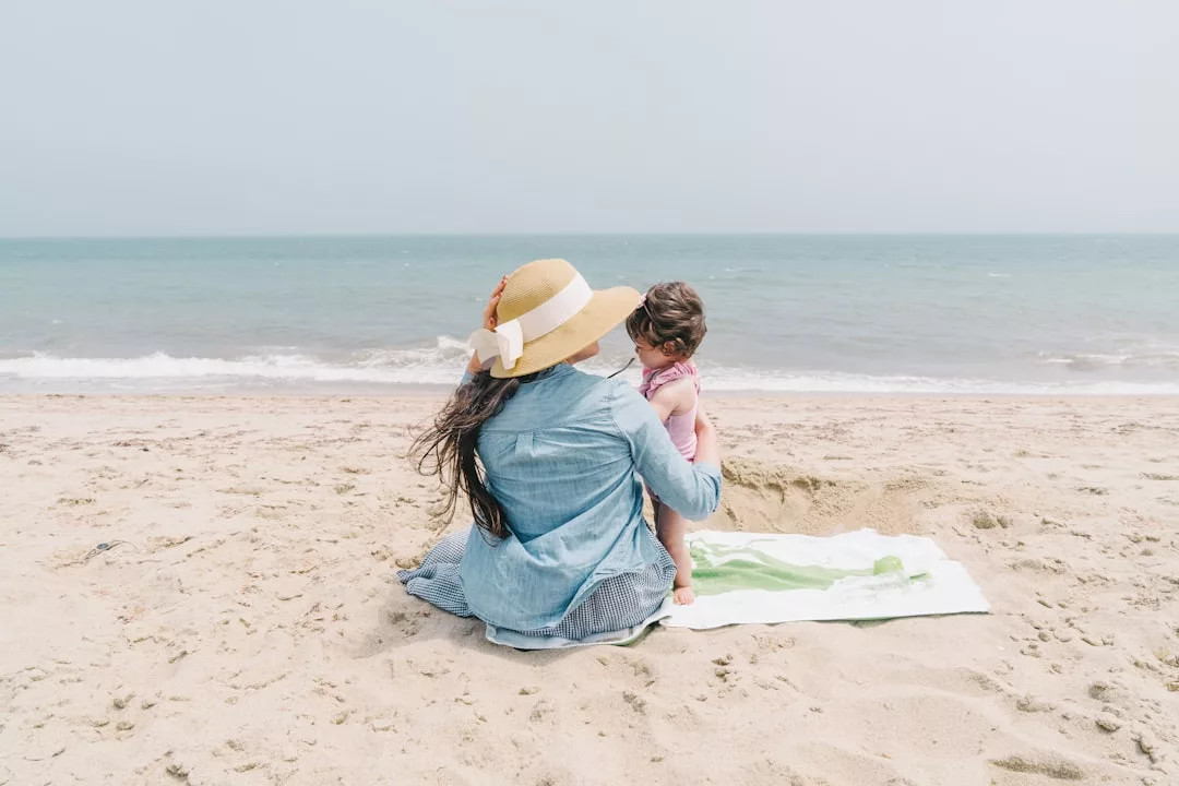 Eine Frau mit Sonnenhut sitzt mit einem kleinen Kind auf einem Strandtuch an einem Sandstrand auf Mallorca unter klarem Himmel und blickt auf das Meer.