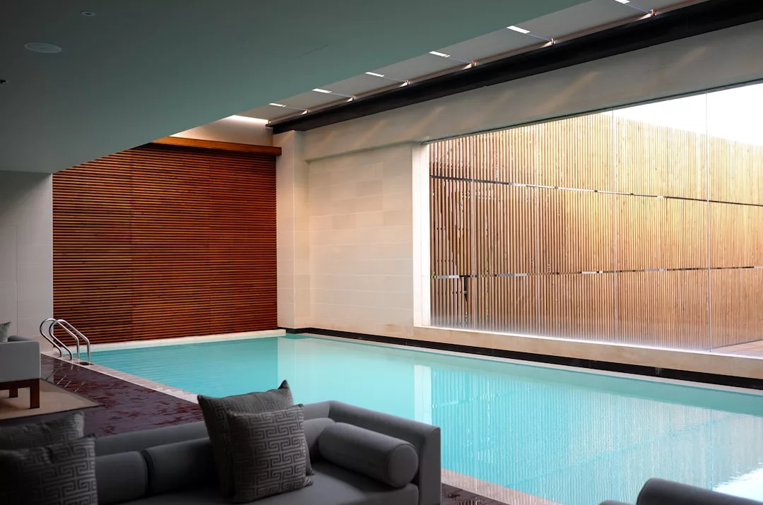 Ein moderner Innenpool mit klarem, blauem Wasser, umgeben von weißen Wänden und Holzlatten, erinnert an die entspannte Eleganz Mallorcas. Im Vordergrund befindet sich eine moderne Sitzecke mit grauen Kissen.