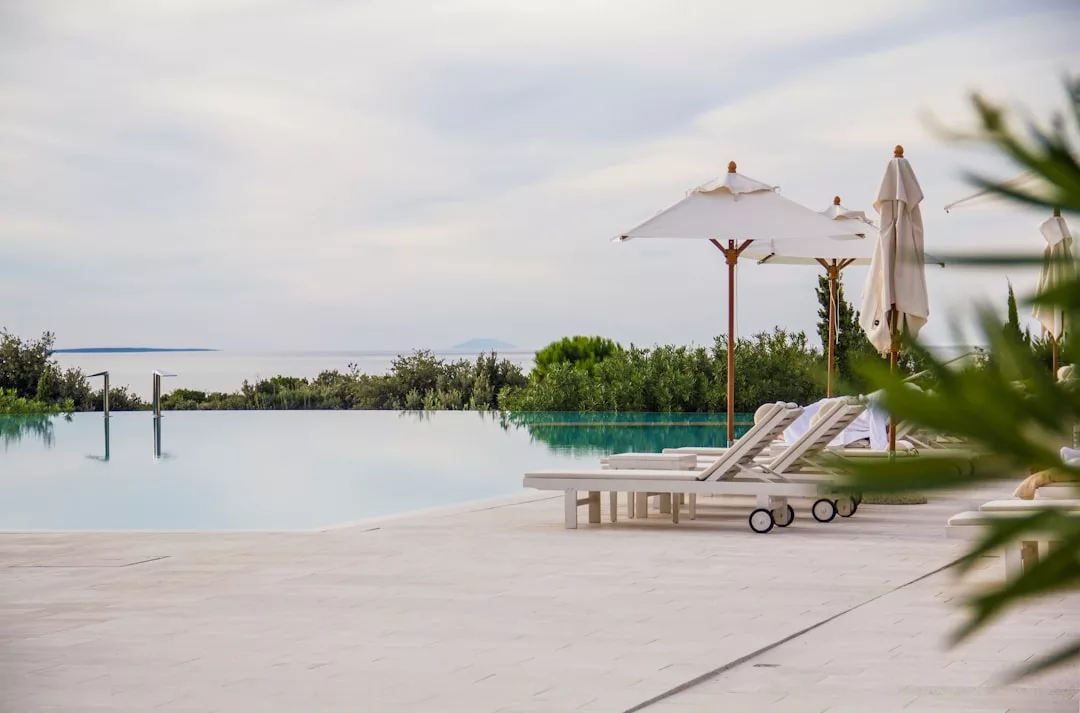 Infinity-Pool mit Liegestühlen und Sonnenschirmen auf einem gefliesten Deck mit Blick auf Mallorcas üppiges Grün und ein Gewässer in der Ferne unter einem bewölkten Himmel.