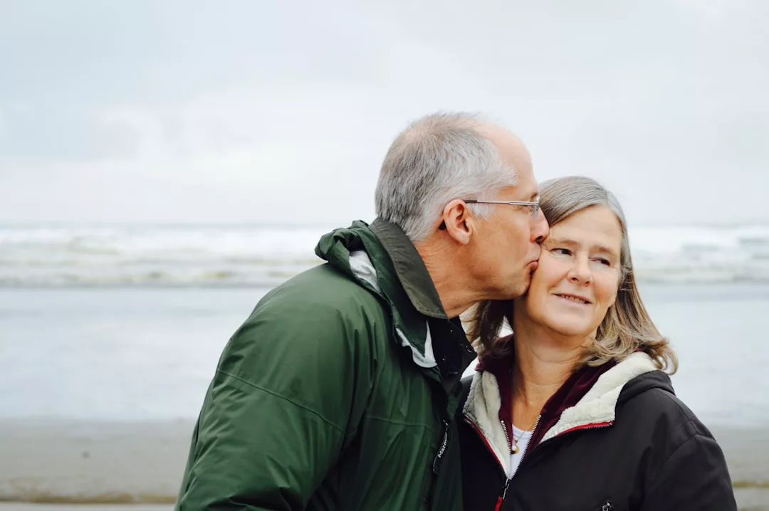 Ein älterer Mann küsst eine Frau auf die Wange. Beide stehen zusammen an einem Strand auf Mallorca. Sie tragen Jacken. Im Hintergrund sind das Meer und der bewölkte Himmel zu sehen.