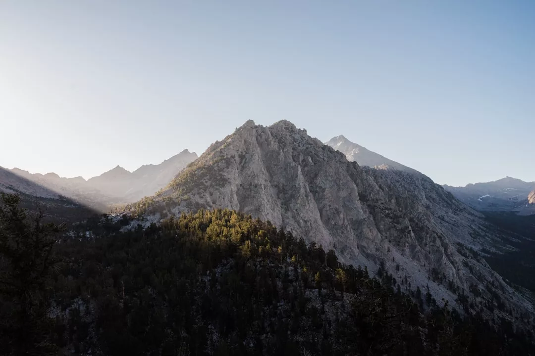 Felsiger Berggipfel auf Mallorca, Sonnenlicht wirft Schatten auf einen bewaldeten Hang unter einem klaren blauen Himmel.