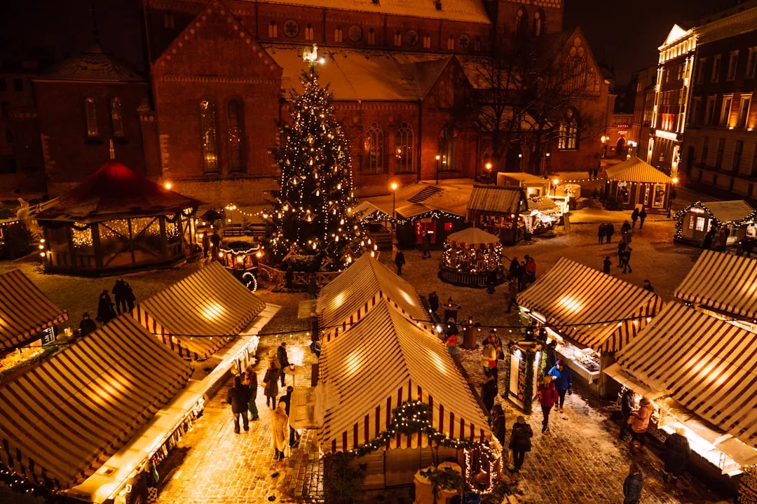 Nachtansicht eines festlichen Weihnachtsmarkts im Freien auf Mallorca mit Ständen mit gestreiften Dächern, flanierenden Menschen und einem großen geschmückten Baum in der Mitte, umgeben von historischen Gebäuden.