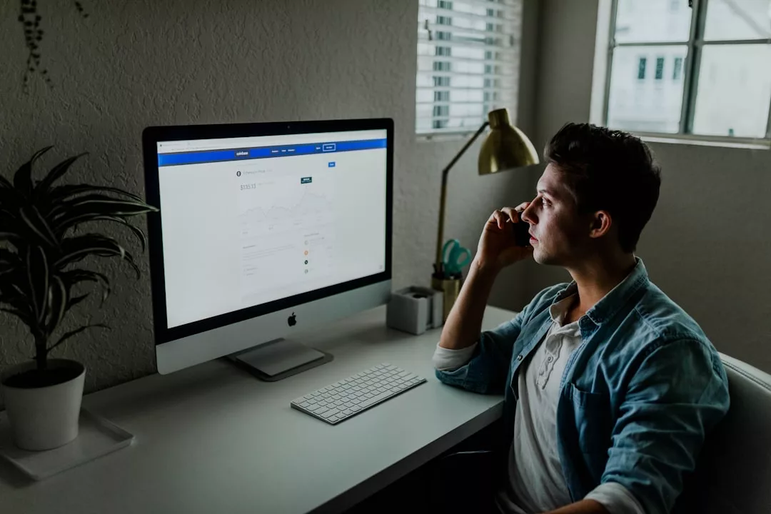 Ein Mann sitzt an einem Schreibtisch in seinem Homeoffice auf Mallorca, schaut auf einen Computerbildschirm und telefoniert. In der Nähe stehen eine Lampe und eine Pflanze.