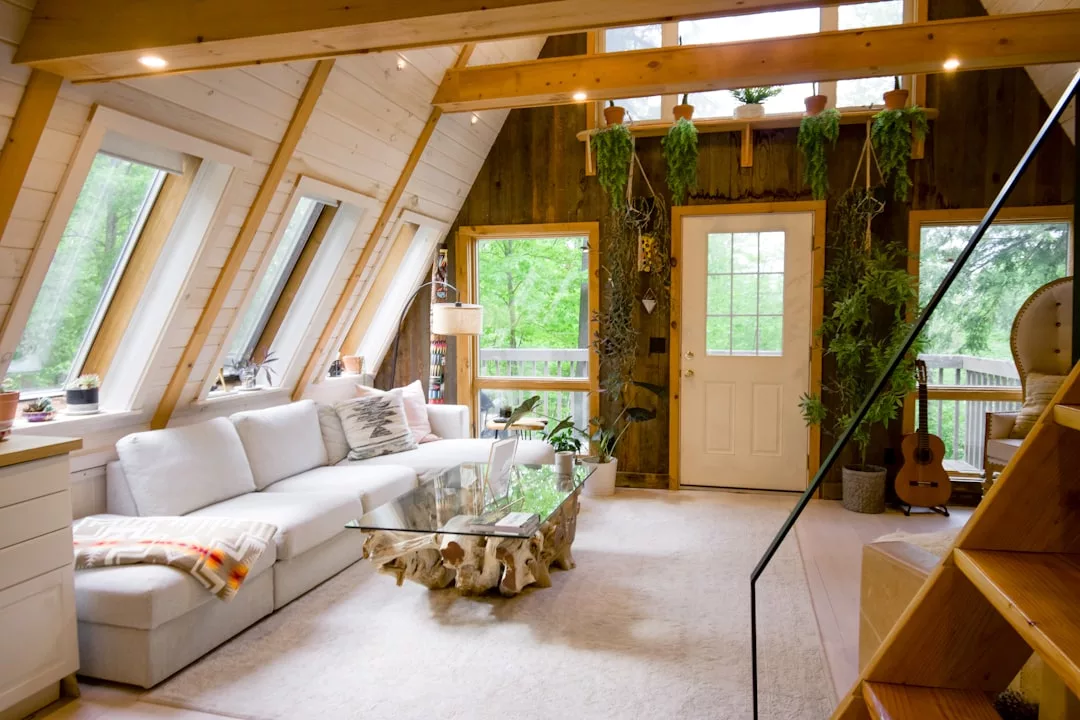 Ein gemütliches Wohnzimmer in A-Form mit großen Fenstern, einem weißen Sofa, einem Couchtisch aus Glas, Hängepflanzen, Holzakzenten und einer Gitarre neben der Tür erinnert an die entspannte Atmosphäre Mallorcas.