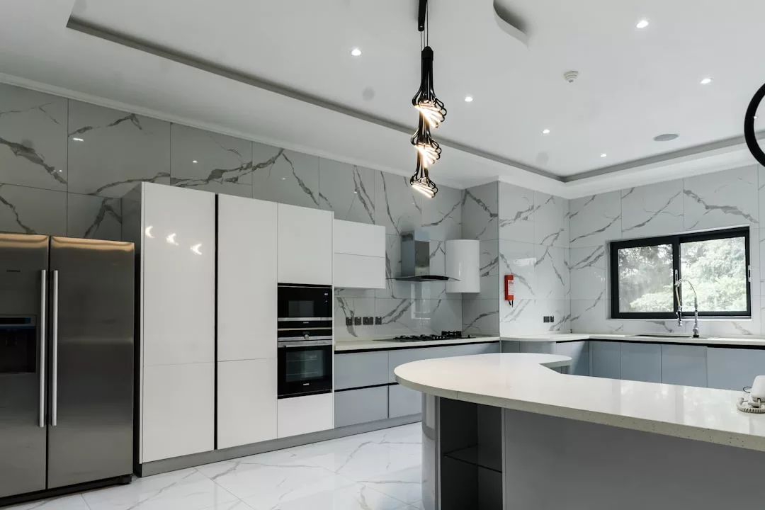 Moderne mallorquinische Küche mit Marmorwänden und -boden, Edelstahlkühlschrank, eingebauten Öfen, Gaskochfeld, weißen Schränken und einer Kochinsel mit Hängelampen.
