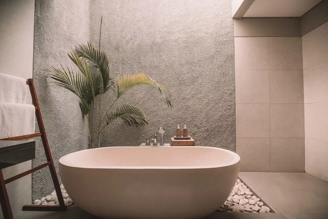 Modernes Badezimmer im Mallorca-Stil mit freistehender ovaler Badewanne, Handtuchhalter, Topfpflanze und Toilettenartikeln auf einem Tablett, mit strukturierten Wänden und glattem Steinboden.